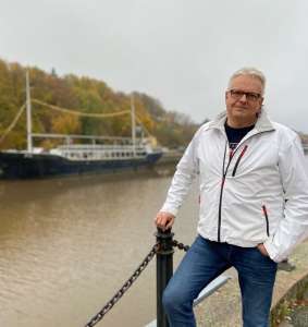 Silmälasipäinen Teemu Nieminen nojaa farkuissa ja valkoisessa takissa tolppaan joen varressa syksyllä, taustalla laiva. 