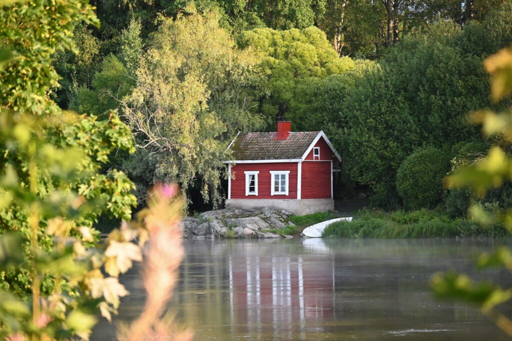 Vihreän metsämaiseman keskellä punainen mökki joen rannalla.