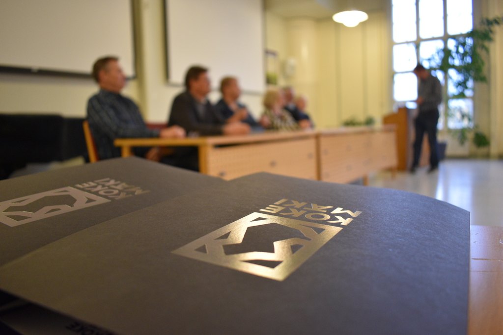 Etualalla musta kirjan kansi, jonka päällä Kokemäen logo, taustalla epäselvänä ihmishahmoja istumassa pöydän ääressä.