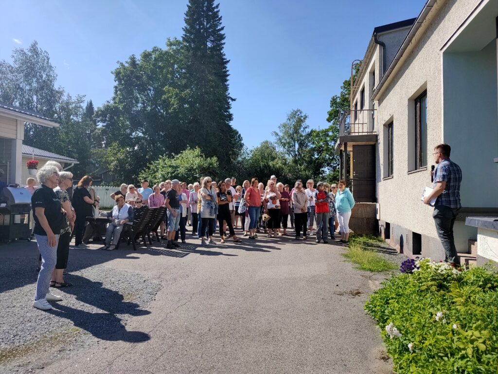 Kuvan oikeassa reunassa vaalea kivitalo, jonka edessä asfaltilla värikkäästi pukeutuneita ihmisiä, auringonpaisteessa. Ihmiset katsovat kameraan. Taustalla vehreitä puita ja sininen taivas.