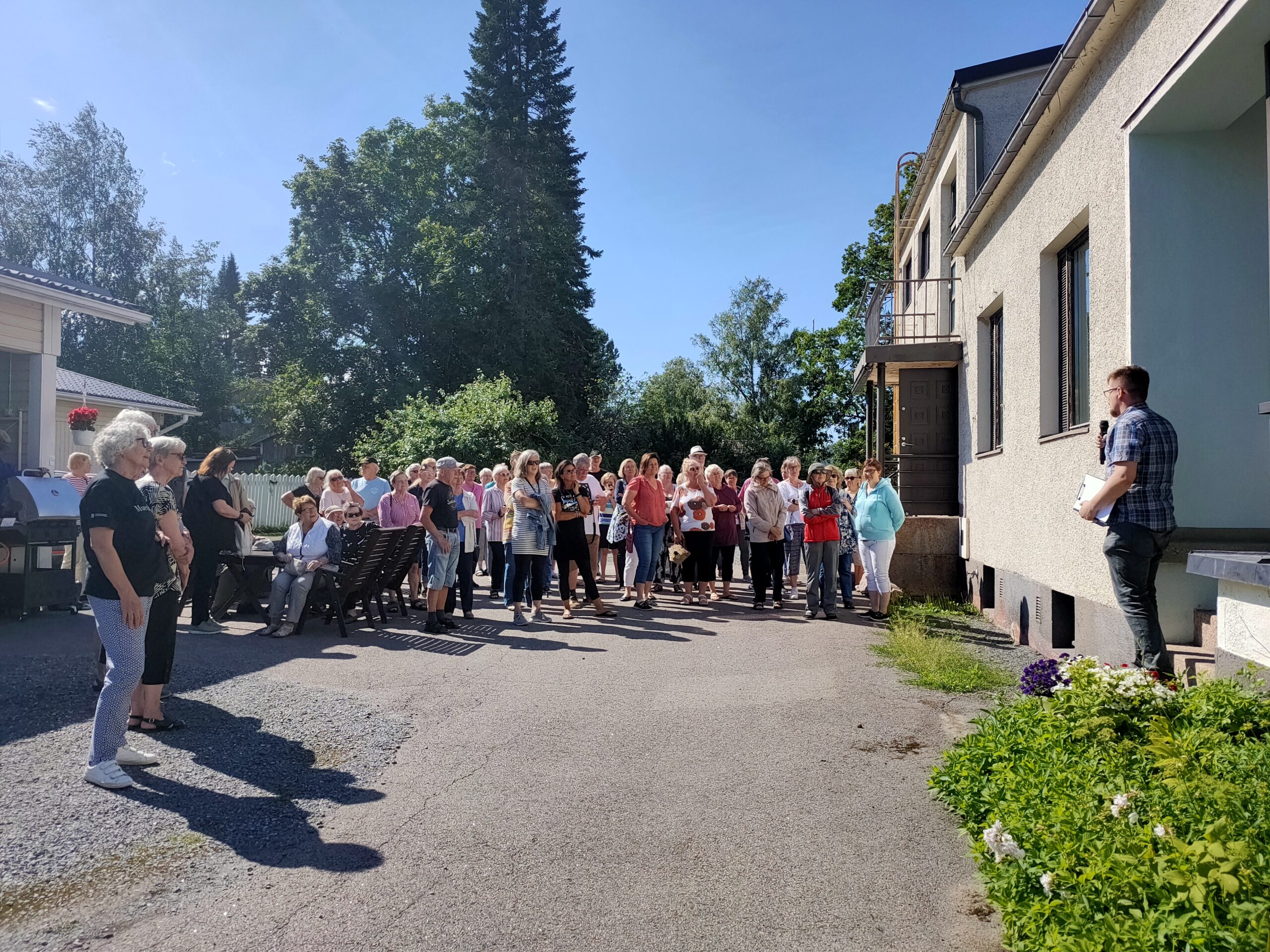 Kuvan oikeassa reunassa vaalea kivitalo, jonka edessä asfaltilla värikkäästi pukeutuneita ihmisiä, auringonpaisteessa. Ihmiset katsovat kameraan. Taustalla vehreitä puita ja sininen taivas.