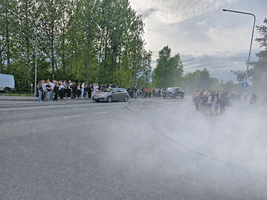 Kadun reunassa ihmisiä, kadulla jarrutusjälkiä ja savua.