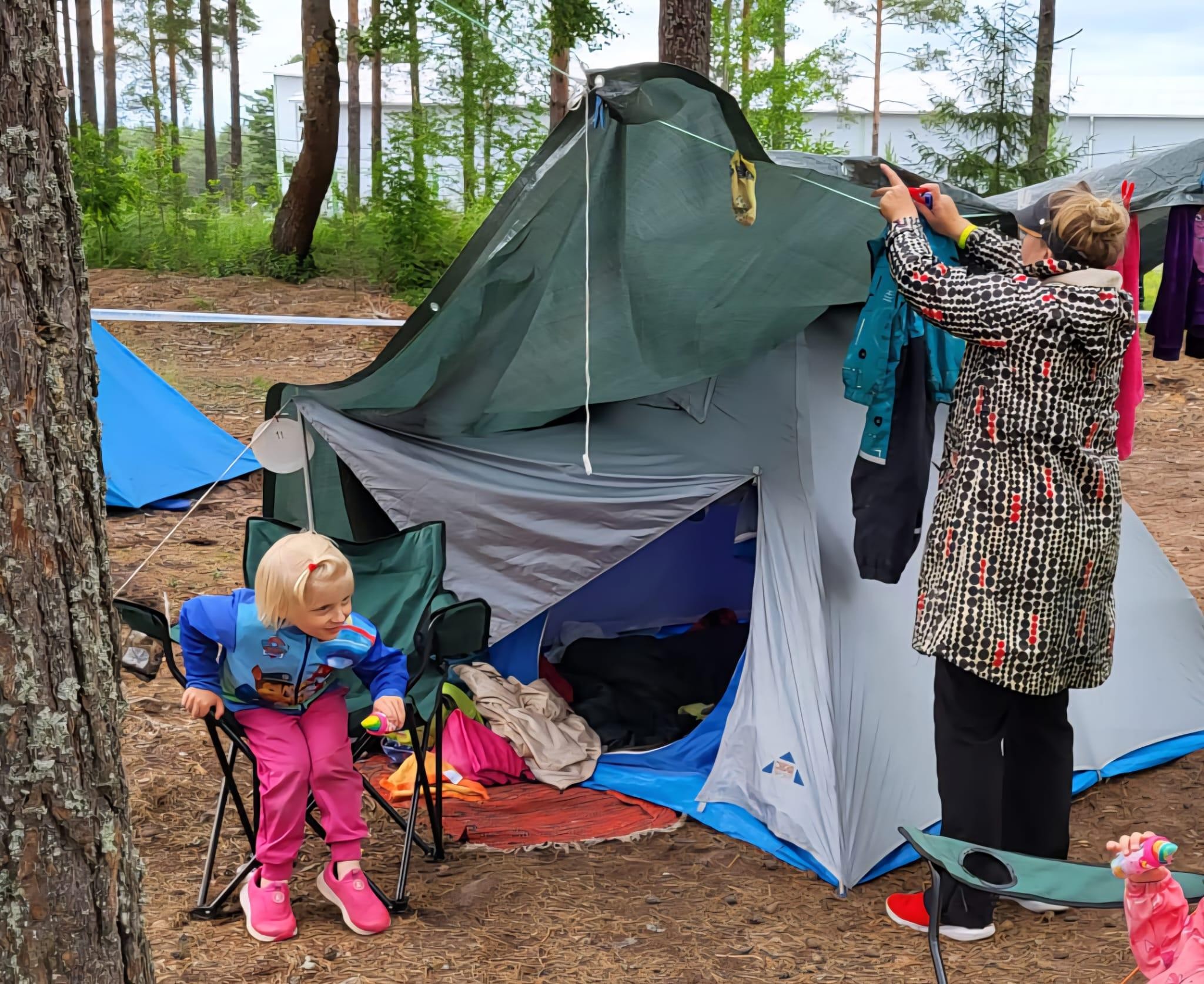 Kuvassa harmaa teltta. Sadettakkiin pukeutunut nainen ripustaa pyykkejä mäntyjen väliin pingotettuun nauhaan. Etualalla lapsi levittelee käsiä sadeasussa.
