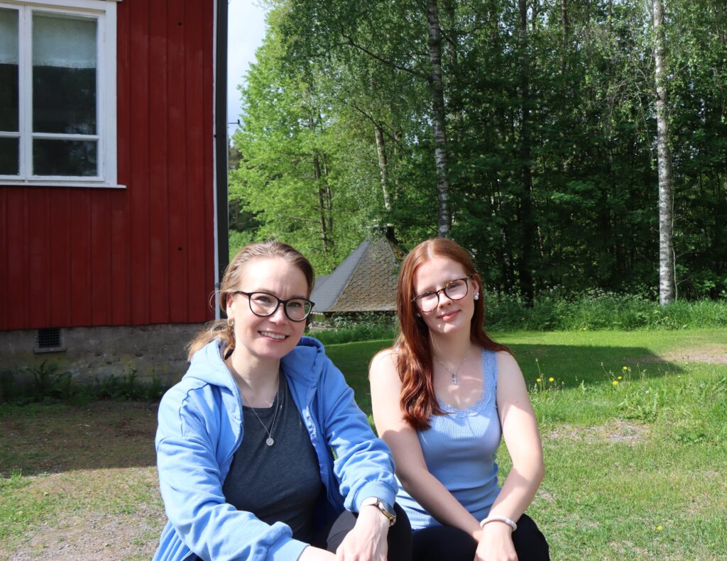 Kesätyöntekijät istumassa kuva etualalla. Taustalla punainen puutalo ja nurmikkoa.