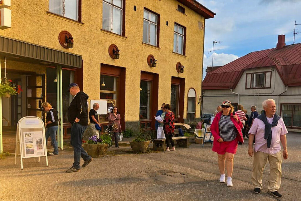 Keltaimem kivitalo vasemmalla, sininen taivas ja ihmisiä edustalla.