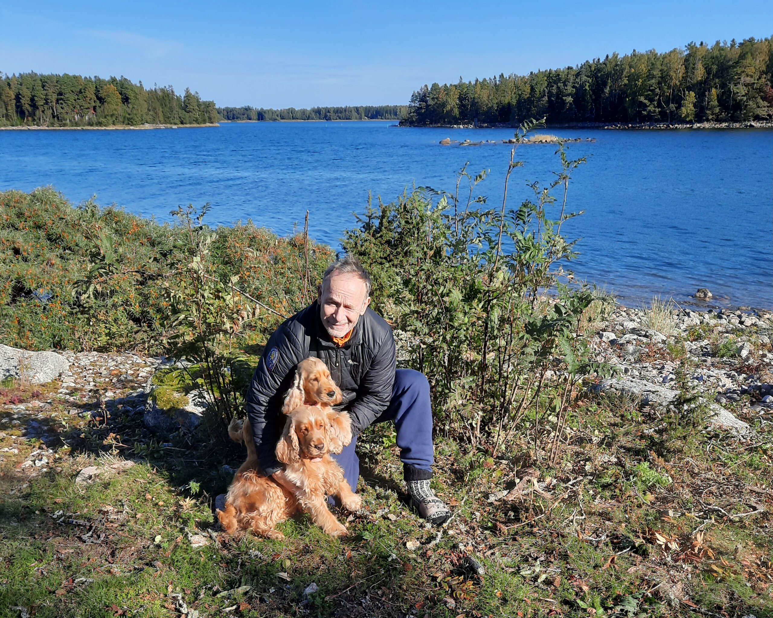 Mies kallioisessa rantamaisemassa. Takana kitukasvuinen pensas. Takana näkyy järvi. Mies hymyilee ja on kyykyssä kahden ruskean koiransa kanssa. Hänellä on päällään ulkoiluvarusteet.