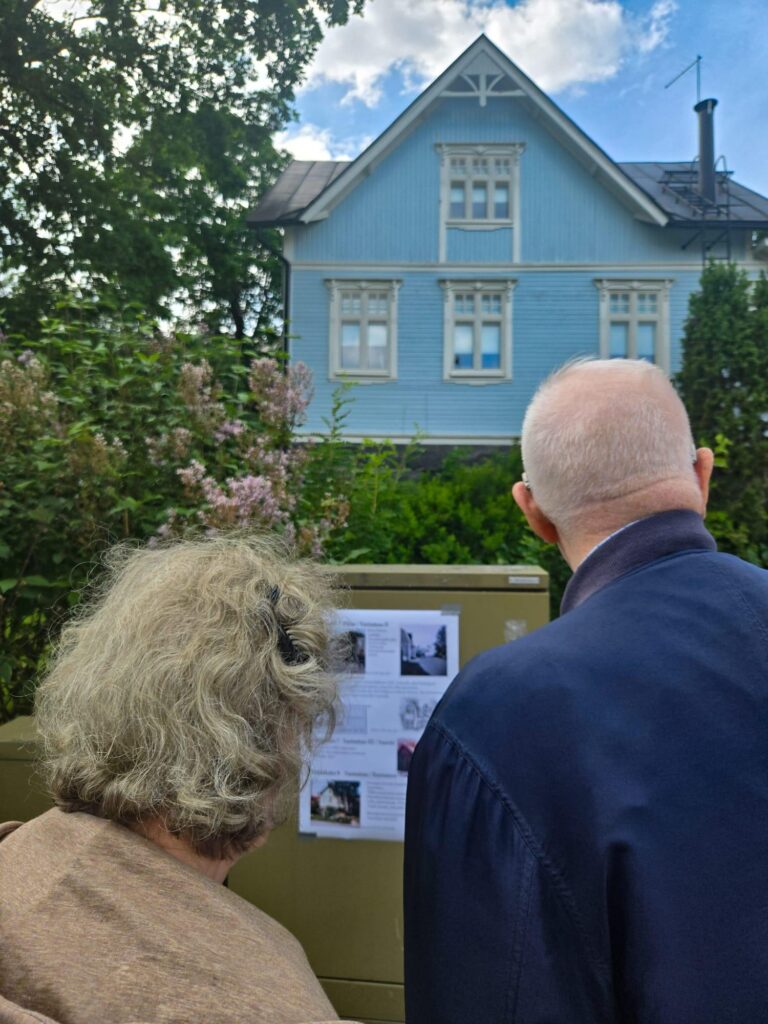Harmaahapsiset vanha nainen ja mies selin kameraan, katsovat esitettä ja taustalla harjakattoinen sininen talo.