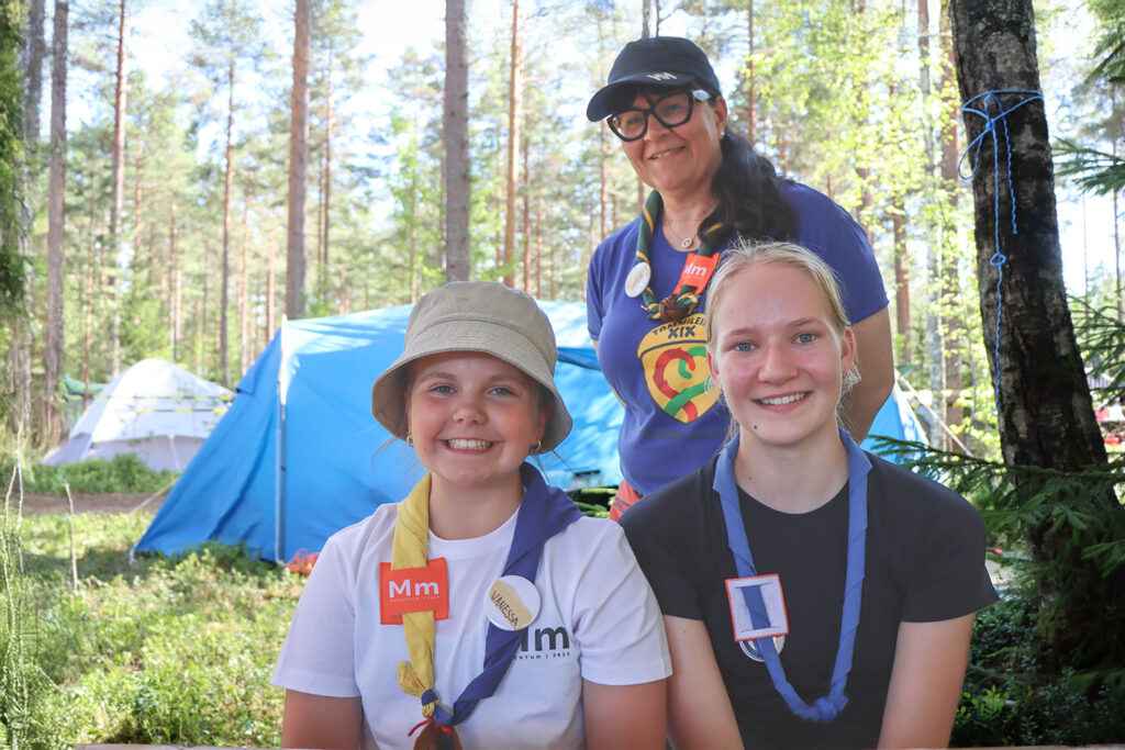 Kolme ihmistä oikealla etualalla taustalla metsä ja teltta.