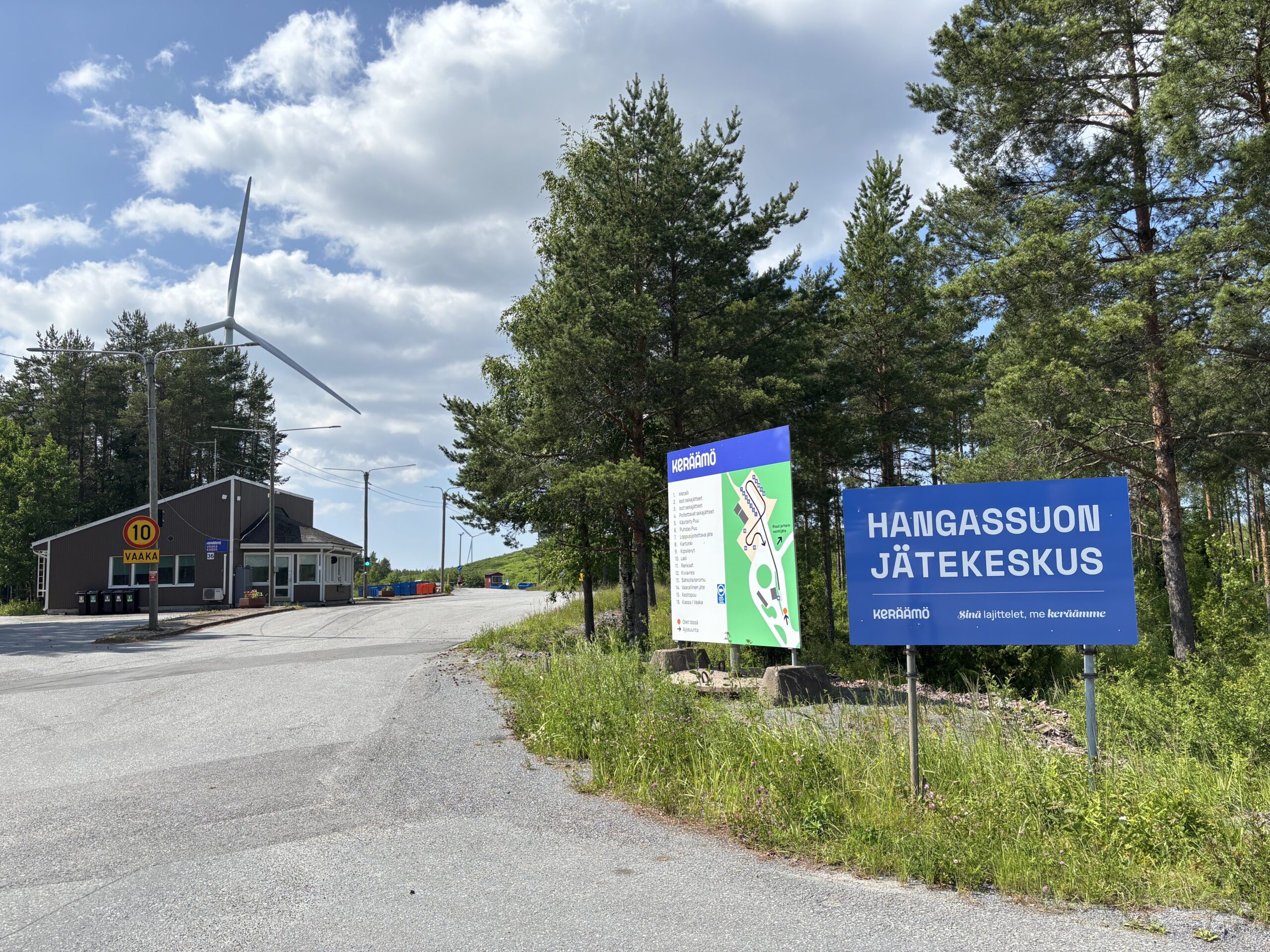 Kuvassa Hangassuon jätekeskus-kyltti, joka on sininen. Vieressä alueen kartta. Taustalla näkyy jätekeskusta ja puita. Takana on tuulivoimala.