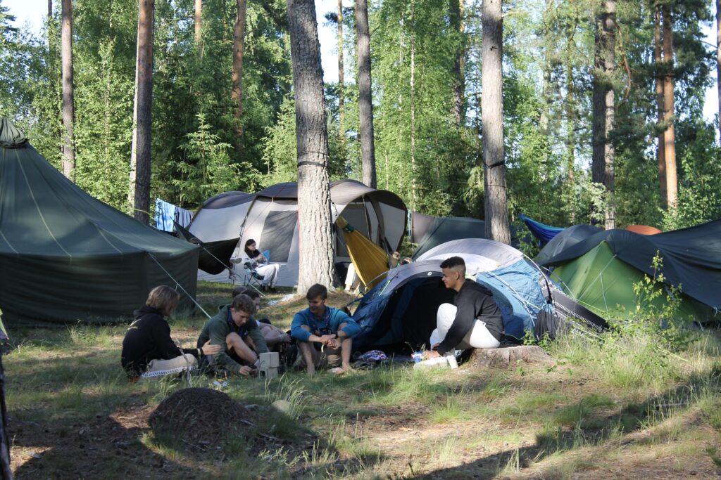 Sini-harmaita telttoja mäntymetsässä. Aurinkoinen päivä. Telttojen edessä istuu ihmisiä, jota juttelevat keskenään rennon oloisesti.
