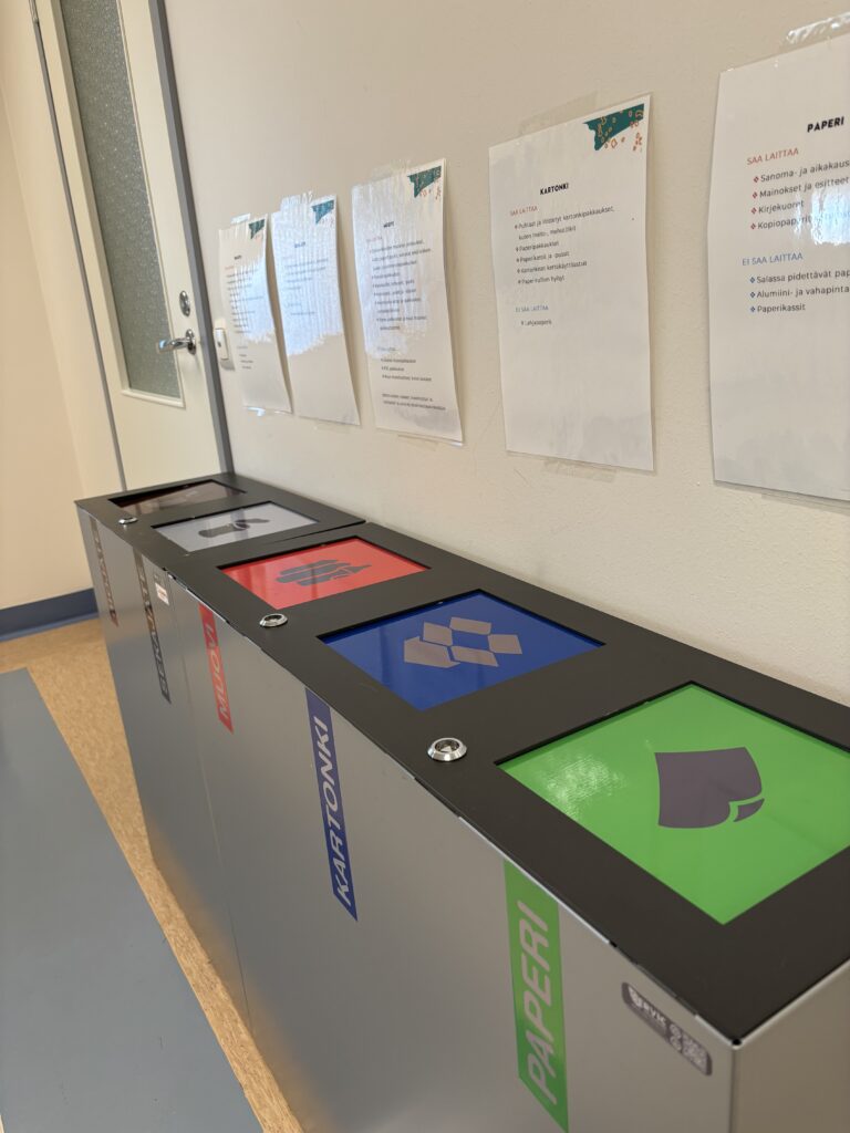 Lajitteluseinämä, joka on harmaa ja noin puolen metrin korkuinen. Seinämässä on erivärisiä luukkuja, joissa on paperi, pahvi, biojäte. metalli ja sekajäte-symbolit