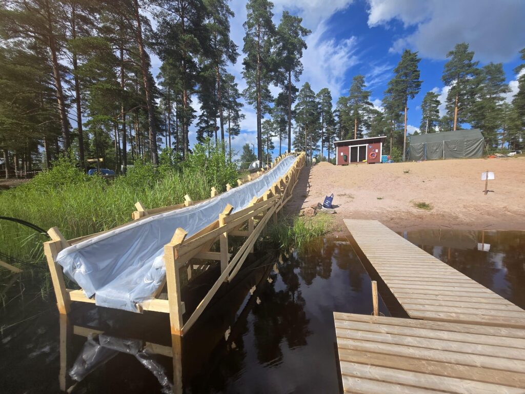 Puita ja sininen taivas, pitkä puusta rakennettu vesiliukumäki laskee Pitkäjärveen.