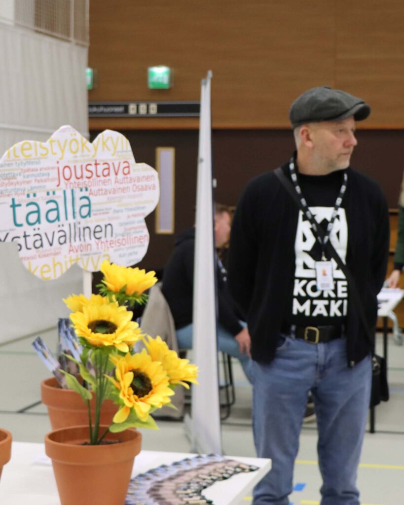 Vasemmalla isot kukat pöydällä, oikealla mies Kokemäki-paita yllä, kuva otettu Tulkkila-salissa.