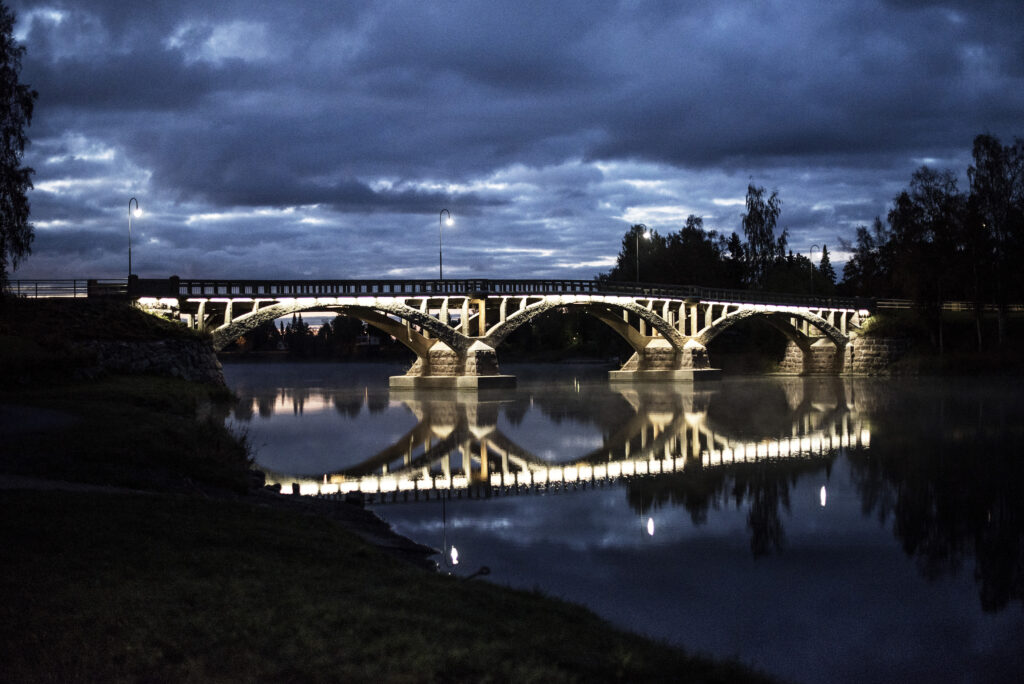 Joki ja silta kuvattuna rannalta. Hämärää, taivas on tummansininen. Silta on valaistu kullanhohtoisesti.